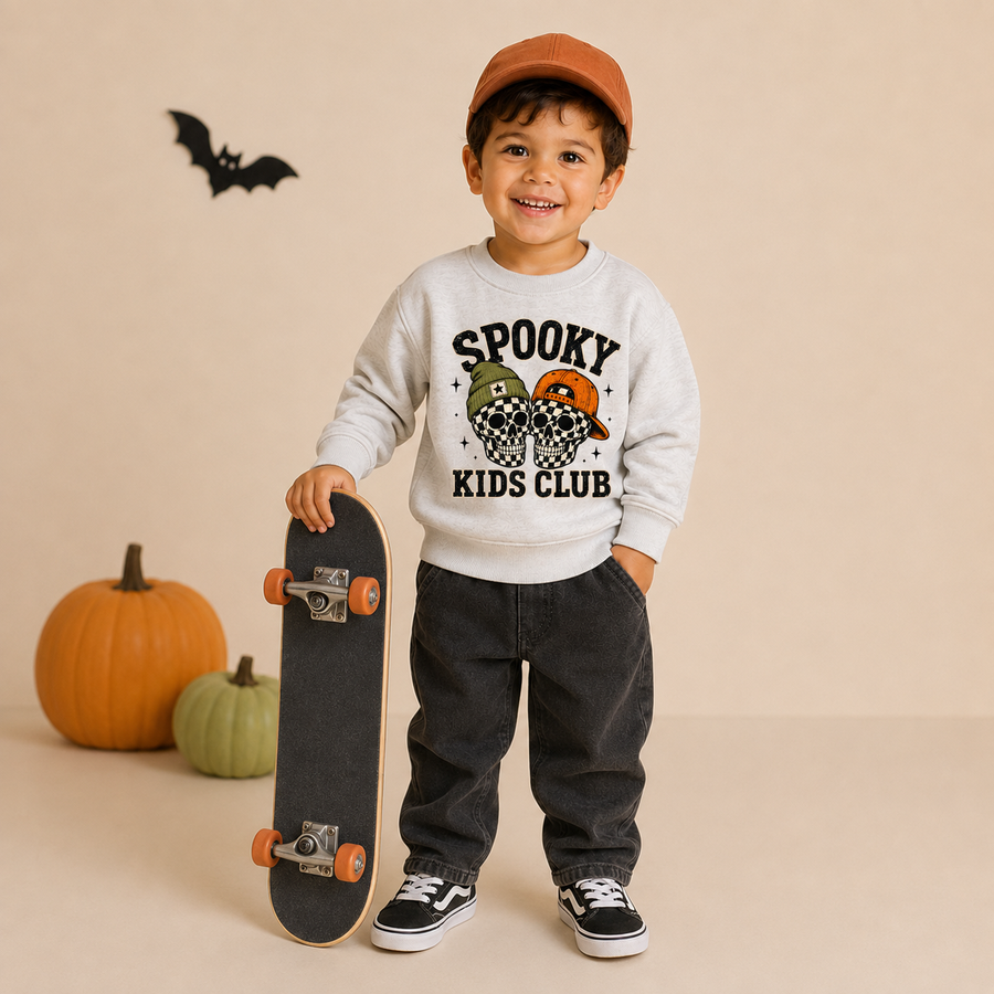 A young boy stands holding a skateboard in a studio setting, wearing a light gray sweatshirt with a 'Spooky Kids Club' graphic, dark gray pants, black sneakers, and an orange cap. Two pumpkins and a black bat decoration are on the floor against a beige background.