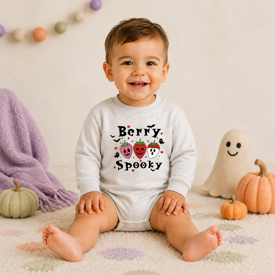 A smiling toddler sits on a patterned rug wearing a light gray long-sleeve bodysuit with a 'Berry Spooky' Halloween graphic. The scene is decorated with small pumpkins, a purple knit blanket, and a ghost toy in a warm, cozy indoor setting.