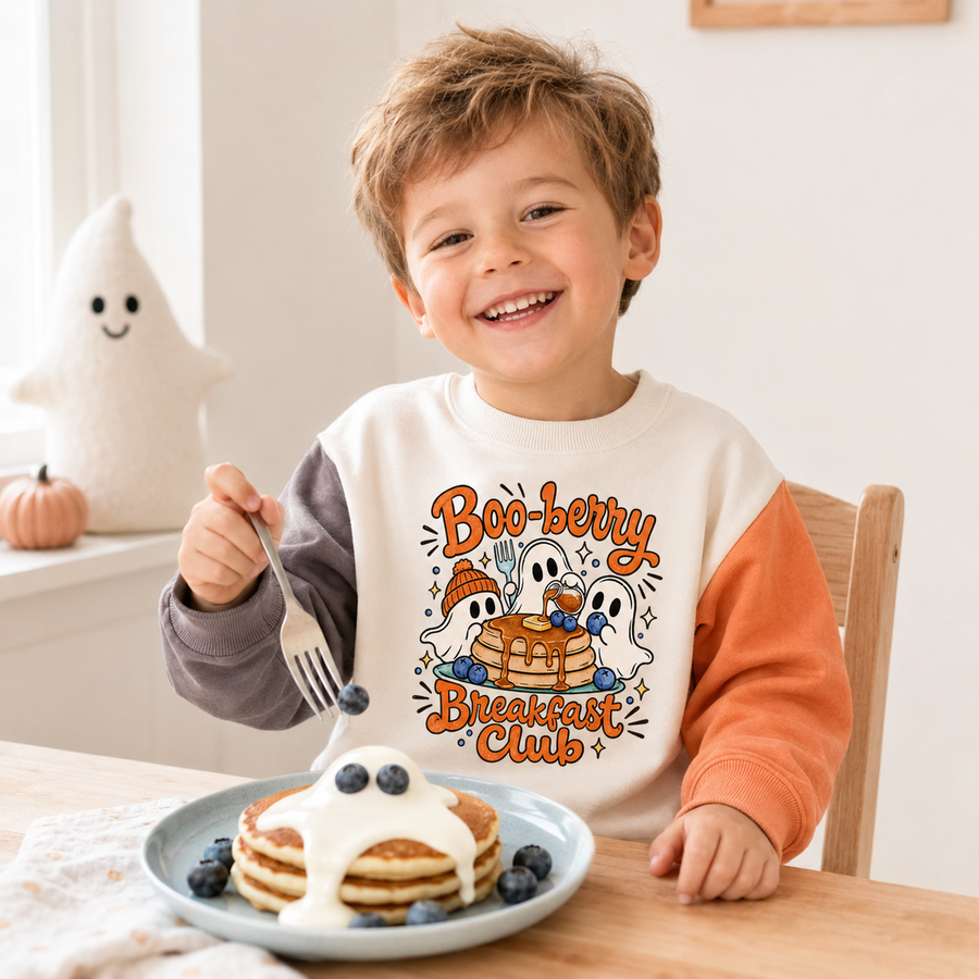 A smiling young boy sits at a wooden table wearing a long-sleeve crewneck sweatshirt with a white body and orange sleeves. The front of the sweatshirt features a colorful graphic of pancakes, blueberries, ghosts, and the text 'Boo-berry Breakfast Club'.