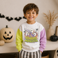 A smiling young boy stands indoors wearing a white sweatshirt with bright yellow-green and lavender sleeves and a colorful Halloween cereal graphic on the front that reads 'Cereal Monster.' The room has a warm neutral wall, pumpkins, dried grass in a vase, and black bat garlands in the background.