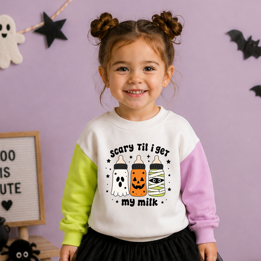 A young girl with twin buns smiles while wearing a white long-sleeve sweatshirt with lime green and lavender color-blocked sleeves. The front of the sweatshirt features a graphic of three baby bottles decorated as a ghost, a jack-o'-lantern, and mummy wrapped in tape, with the text 'scary til i get my milk'.
