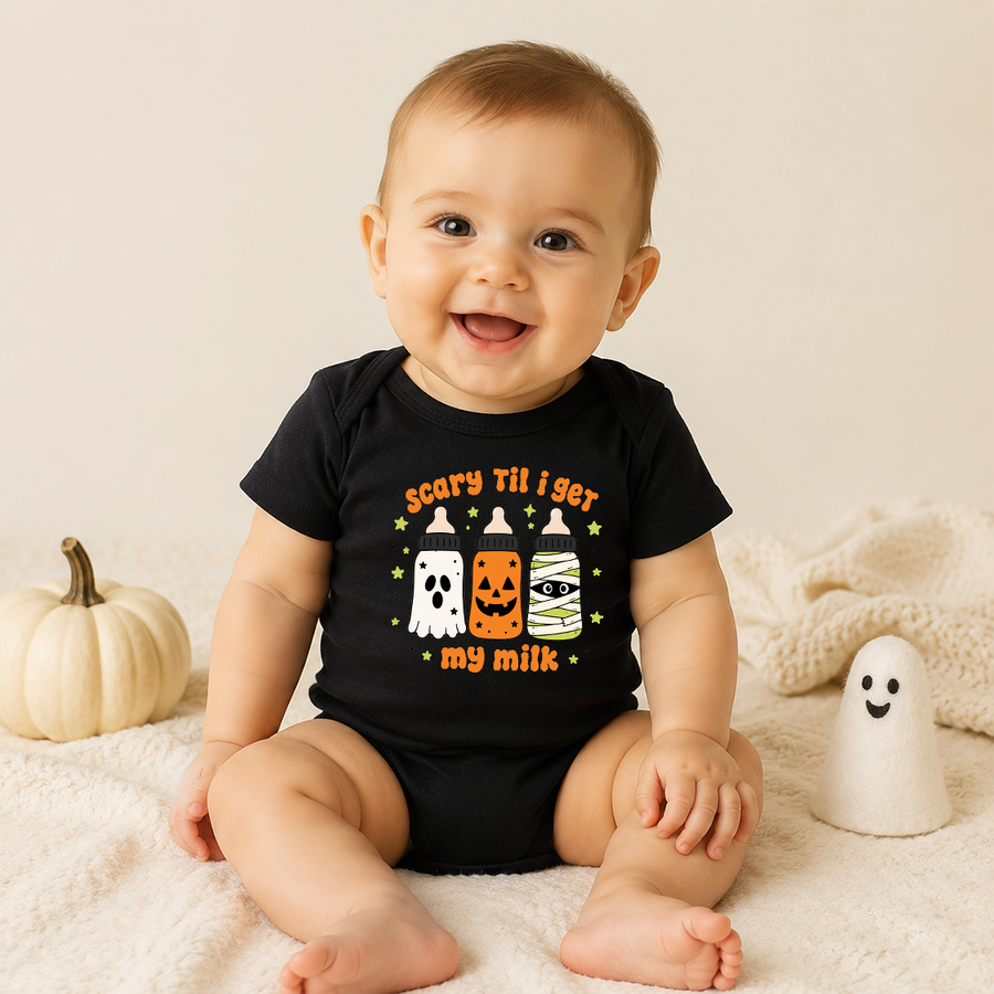 A smiling baby sits on a cream blanket wearing a black short-sleeve bodysuit with a Halloween graphic that reads 'scary til i get my milk' above three cartoon treats and below them. A small white pumpkin and a white ghost toy are beside the baby against a soft beige background.