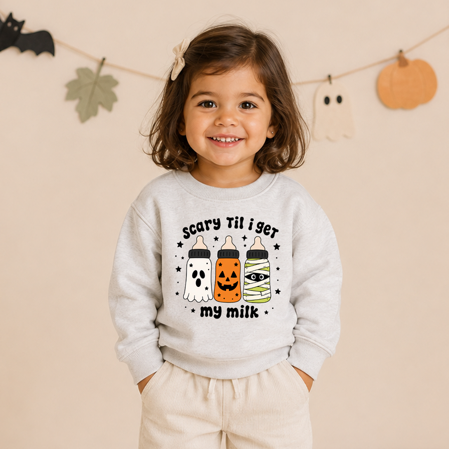 A young child wearing a light gray sweatshirt with a Halloween graphic that reads 'Scary Til i get my milk' above three milk bottles and below them, paired with light beige pants, standing against a warm beige wall with paper decorations.