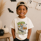 A smiling young boy wearing a beige baseball cap backwards and a cream graphic T-shirt with a skeleton skateboarder and the words 'Skate Slice Repeat!' in a Halloween-themed room with ghost decorations and pumpkins.