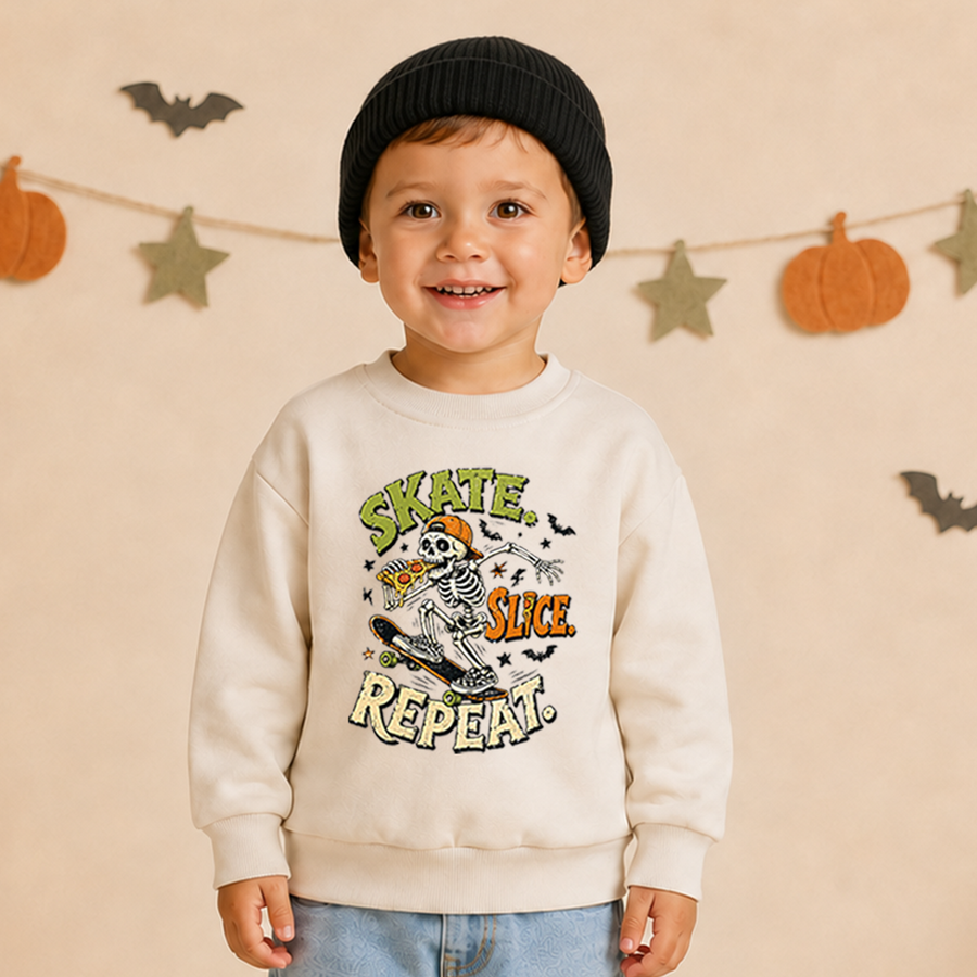 A young child wearing a cream sweatshirt with a colorful 'Skate Slice Repeat' skeleton graphic, paired with a black beanie and light blue jeans, standing against a warm beige background decorated with autumn-themed paper garlands.