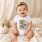 A baby wearing a white short-sleeve bodysuit with a colorful 'Skate, Sue, Repeat!' Halloween graphic, sitting on a cream blanket with decorative pumpkins and a small ghost toy.