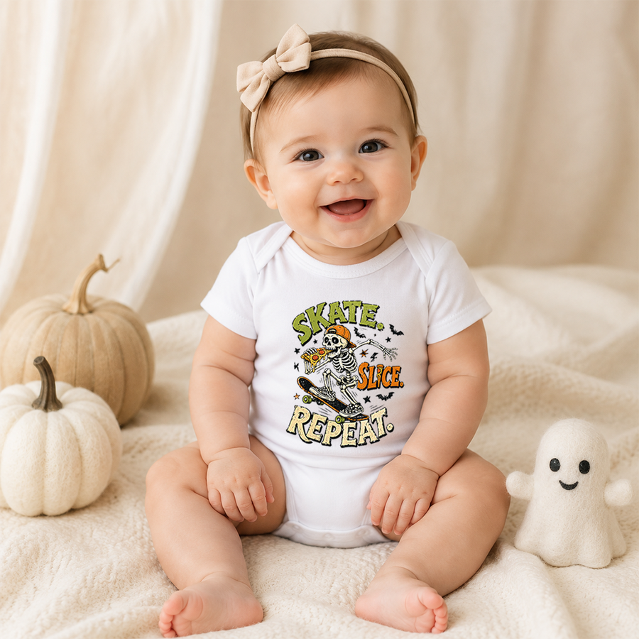 A baby wearing a white short-sleeve bodysuit with a colorful 'Skate, Sue, Repeat!' Halloween graphic, sitting on a cream blanket with decorative pumpkins and a small ghost toy.