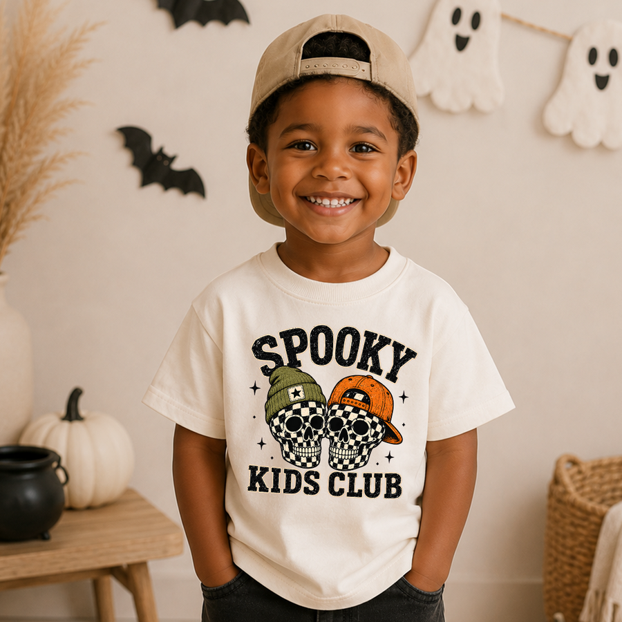 A smiling young boy wearing a beige baseball cap backwards and a cream-colored graphic t-shirt that reads 'Spooky Kids Club' with a skull-and-cap design, standing in a decorated Halloween-themed room.