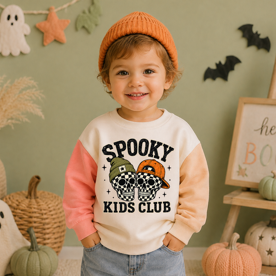 A smiling young child wearing a cream sweatshirt with peach and pink sleeves, blue jeans, and an orange knit beanie. The sweatshirt has a graphic that reads 'SPOOKY KIDS CLUB' with two cartoon hats.