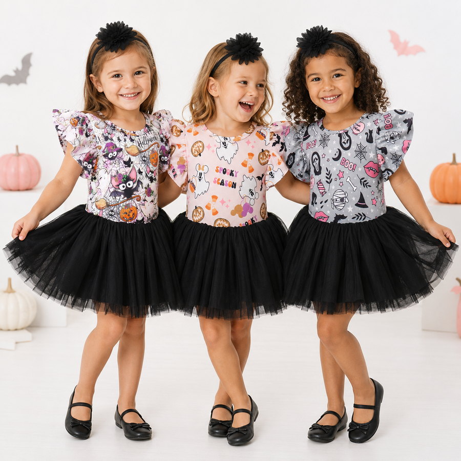 Three young girls standing together and smiling, wearing matching Halloween-themed tops with black tulle tutus and black shoes. The tops feature colorful spooky prints including pumpkins, ghosts, bats, and cats, with one top reading 'SCARY NIGHT'.