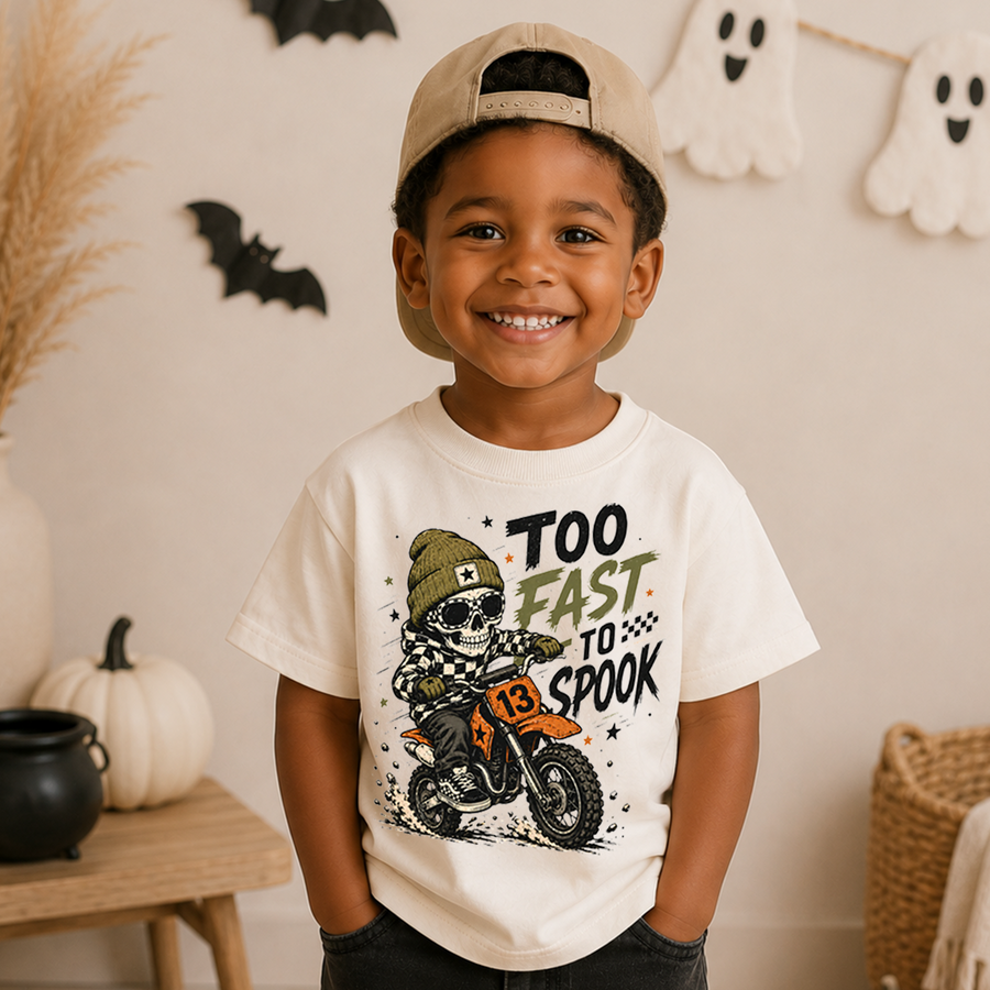 A smiling young boy wearing a cream-colored graphic t-shirt and a beige baseball cap, posed in a cozy, decorated Halloween-themed room with pumpkins, a cauldron, and ghost decorations.