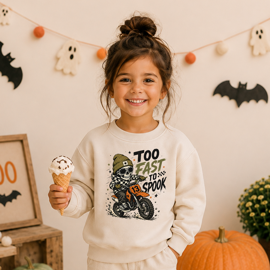 A young girl with her hair in a bun smiles while holding an ice cream cone, wearing a cream-colored sweatshirt with a graphic of a skeleton riding a dirt bike and the text 'TOO FAST TO SPOOK'. The background is decorated for Halloween with ghost and bat cutouts, a pumpkin, and a string of orange and white balloons.
